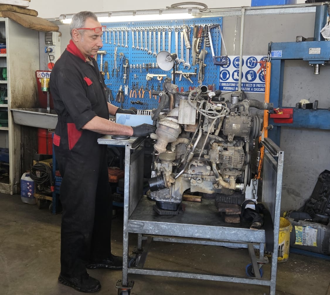 Meccanico in tuta nera e rossa al lavoro su un motore smontato posato su un carrello metallico, davanti a un pannello di attrezzi blu